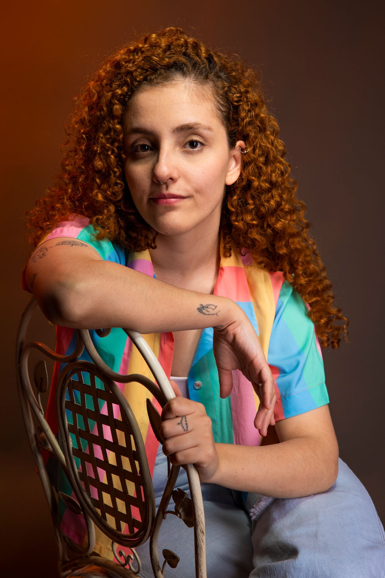 A woman with red curly hair and tattoos sitting on a bentwood chair in warm studio light.