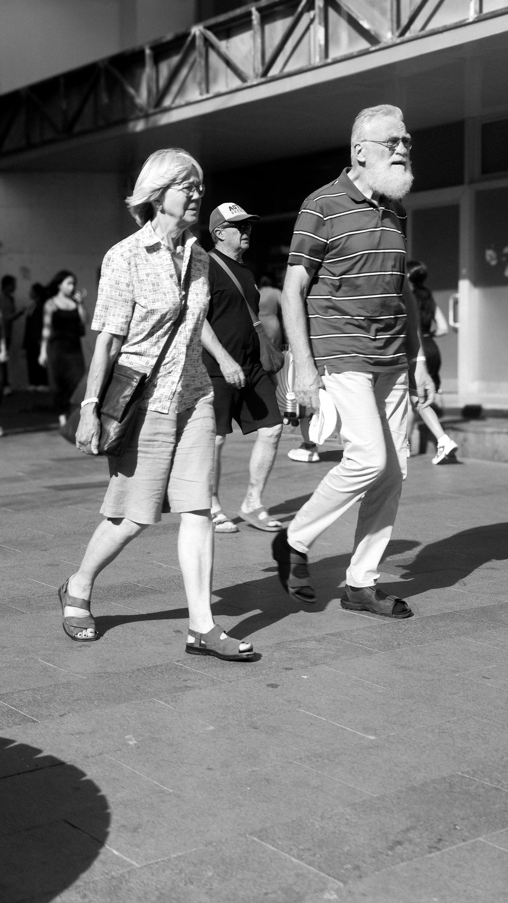 An elderly couple walking side by side through a sunlit plaza, black and white.