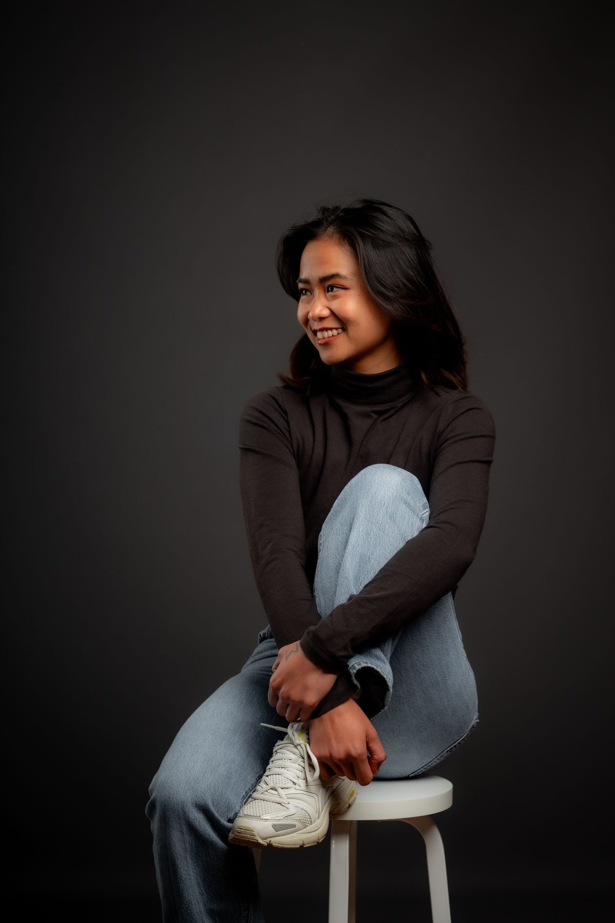 A woman in jeans and a black top sitting on a stool in the studio, looking over her shoulder.