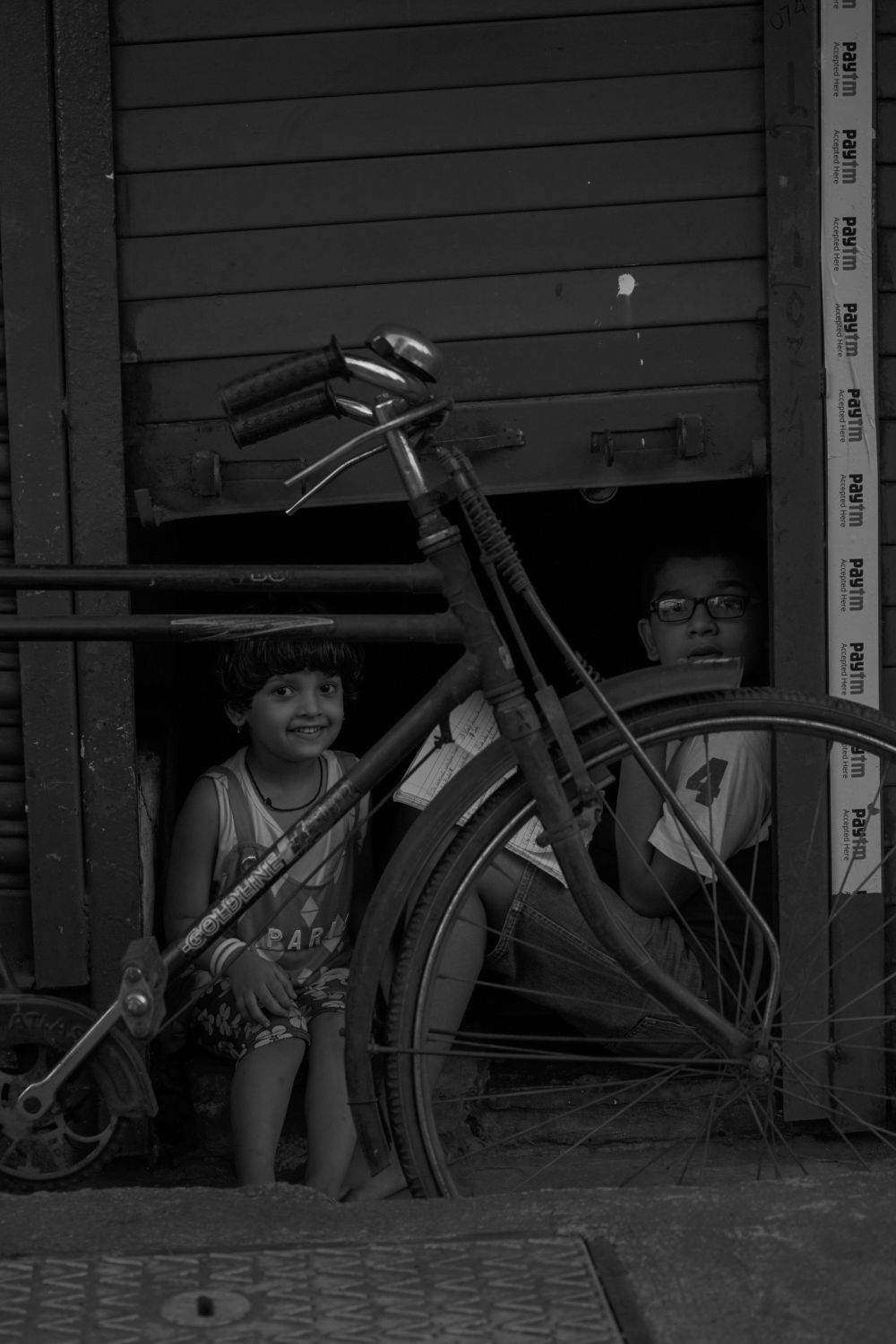 Two kids peeking out from behind a bicycle in a doorway, black and white.