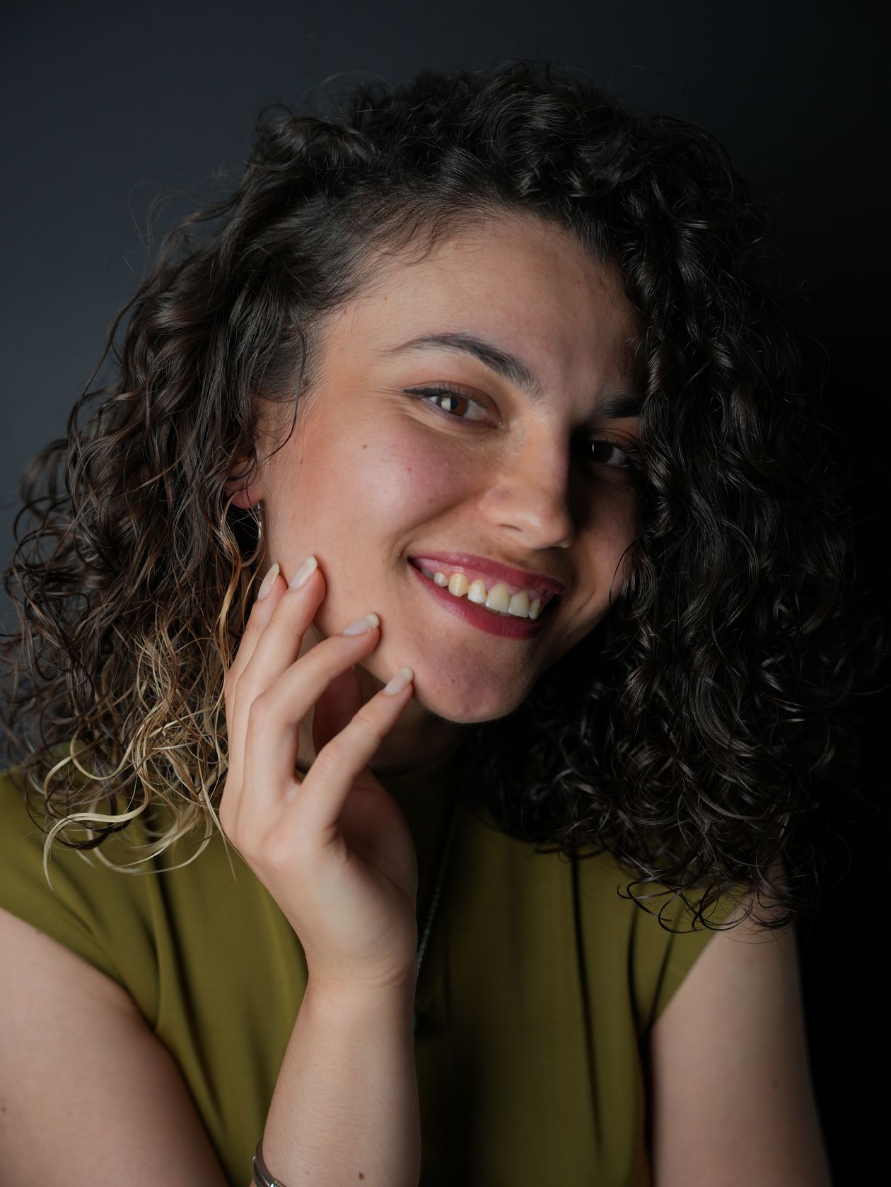 Noa Castellano smiling in a dark studio, curly hair and an olive top.