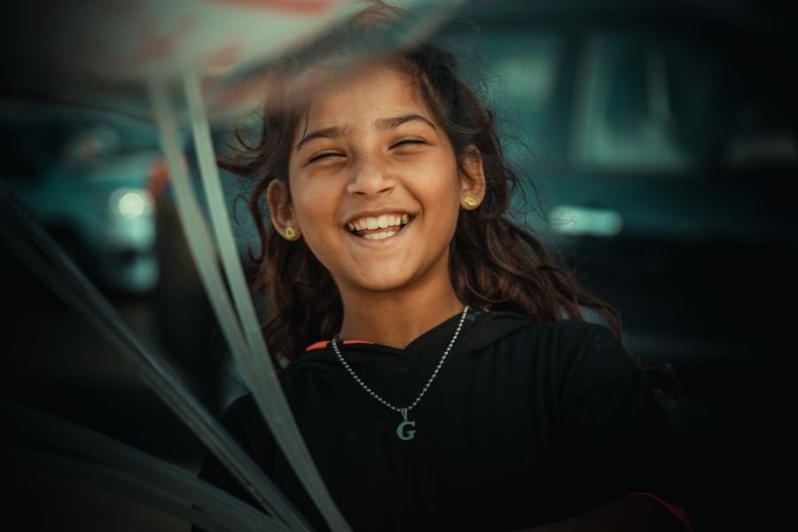 A laughing girl with a beaded necklace, photographed close on a city street.