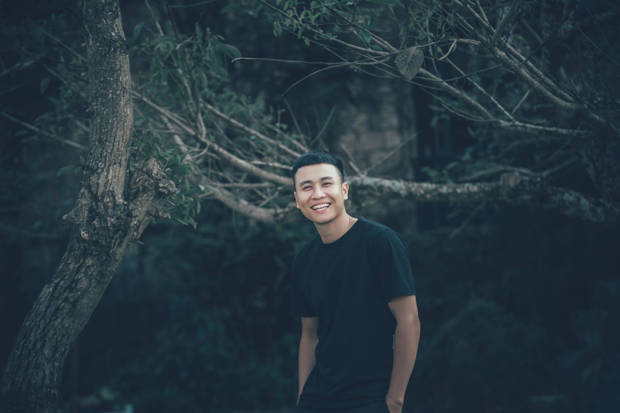 A man in a black t-shirt smiling among trees.
